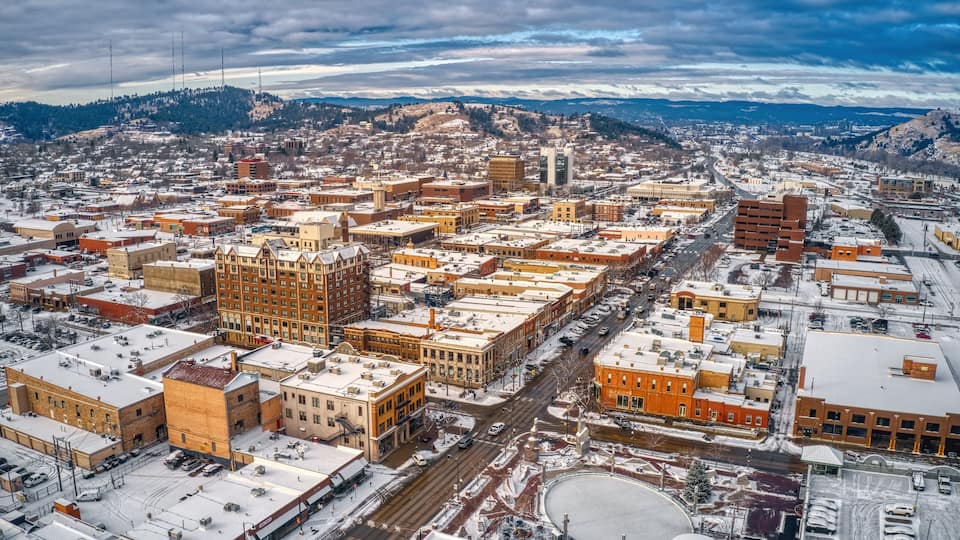 An aerial view of a city with snow-covered buildings and streets, a prominent clock tower, and a hilly backdrop.