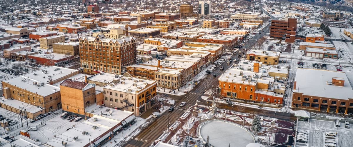An aerial view of a city with snow-covered buildings and streets, a prominent clock tower, and a hilly backdrop.