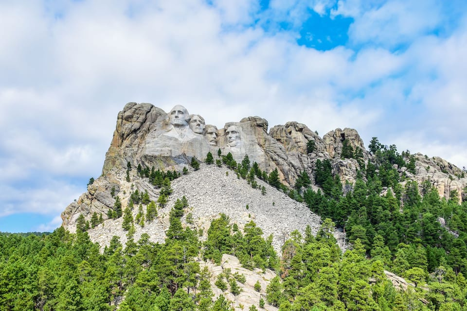 A mountain with a large rock face featuring carved faces, surrounded by dense green forest.