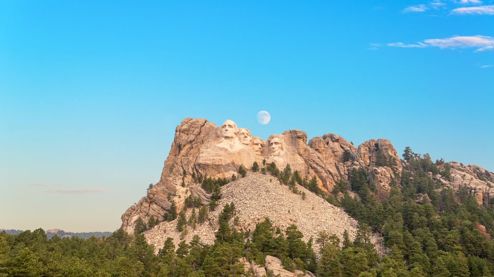 A mountain with a large rock formation resembling a face, a clear sky, and a visible moon.