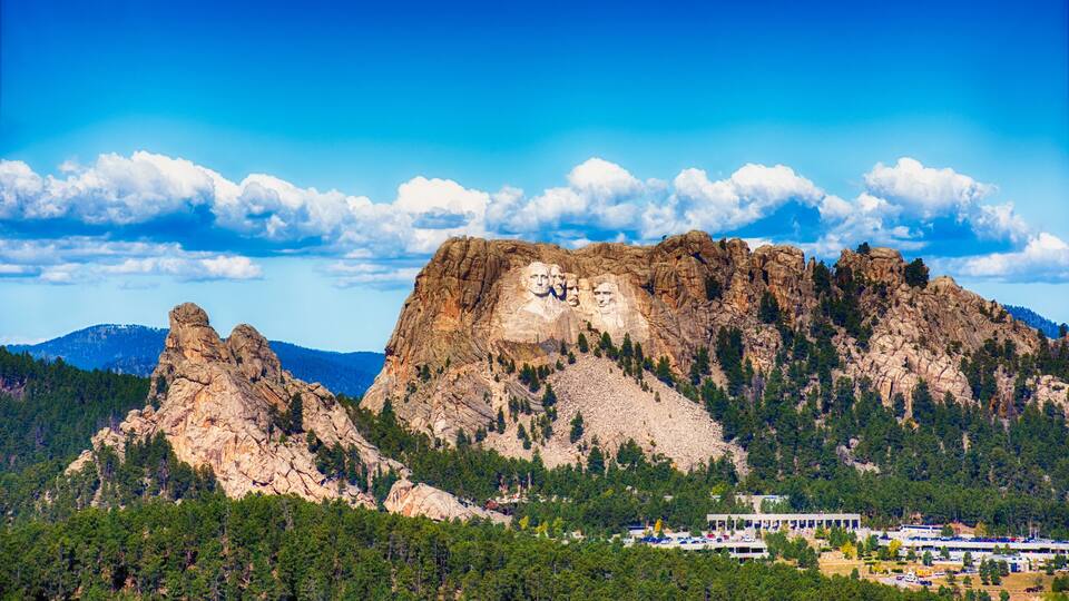 A mountain with carved faces, a clear blue sky, and a forested area with buildings at the base.