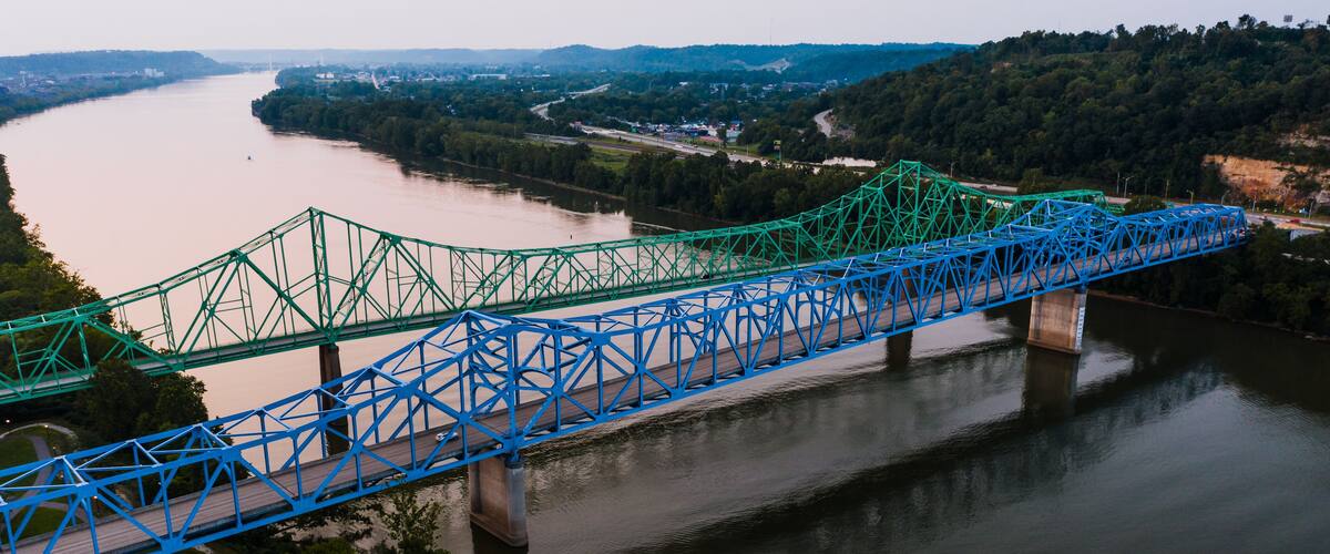 A blue and green bridge spanning a river with surrounding greenery and distant hills.