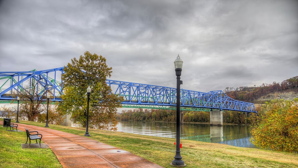 A blue bridge over a river, a park with a paved walkway, and a lamppost.