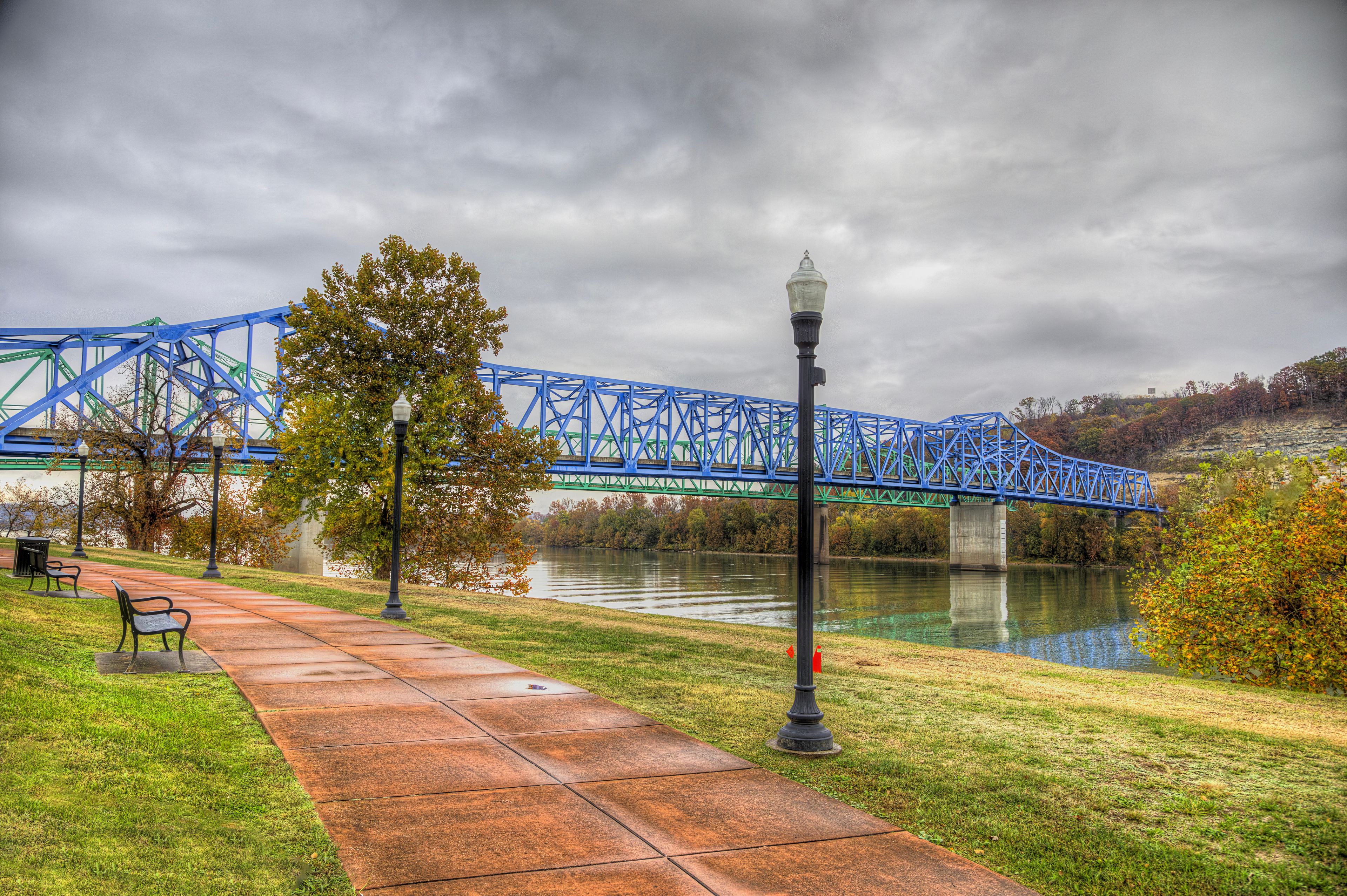 A blue bridge over a river, a park with a paved walkway, and a lamppost.