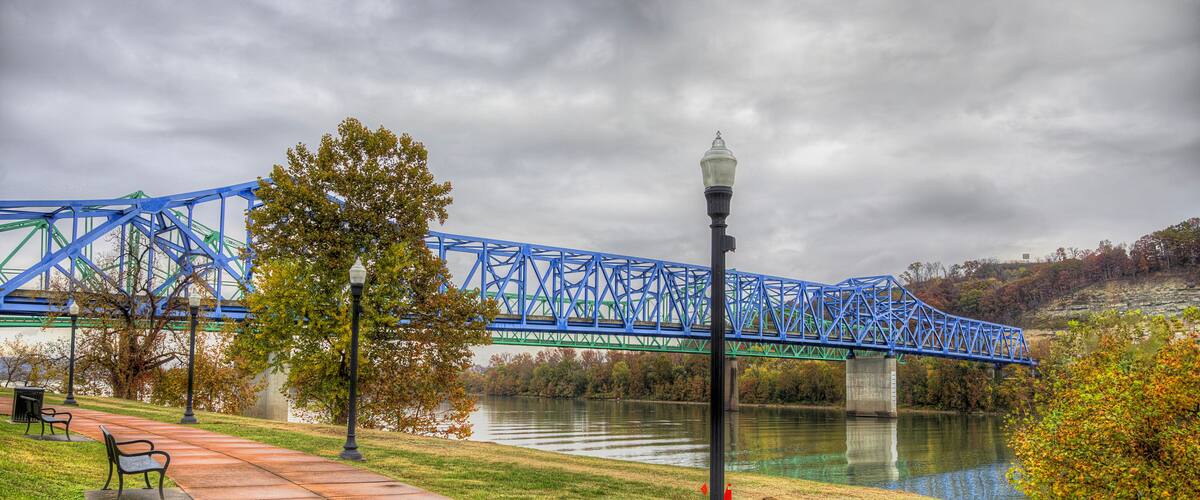 A blue bridge over a river, a park with a paved walkway, and a lamppost.