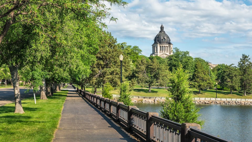 A park pathway lined with trees, a body of water, and a historic building with a dome in the background.
