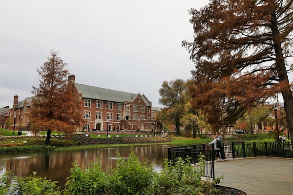 A large brick building with a pond in front, surrounded by trees and a paved walkway.