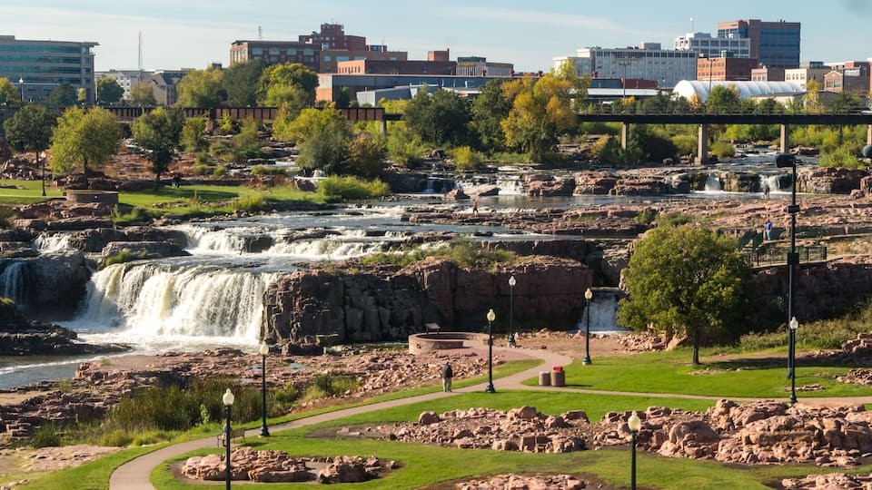 A waterfall cascading over rocky terrain, surrounded by greenery and urban buildings in the background.