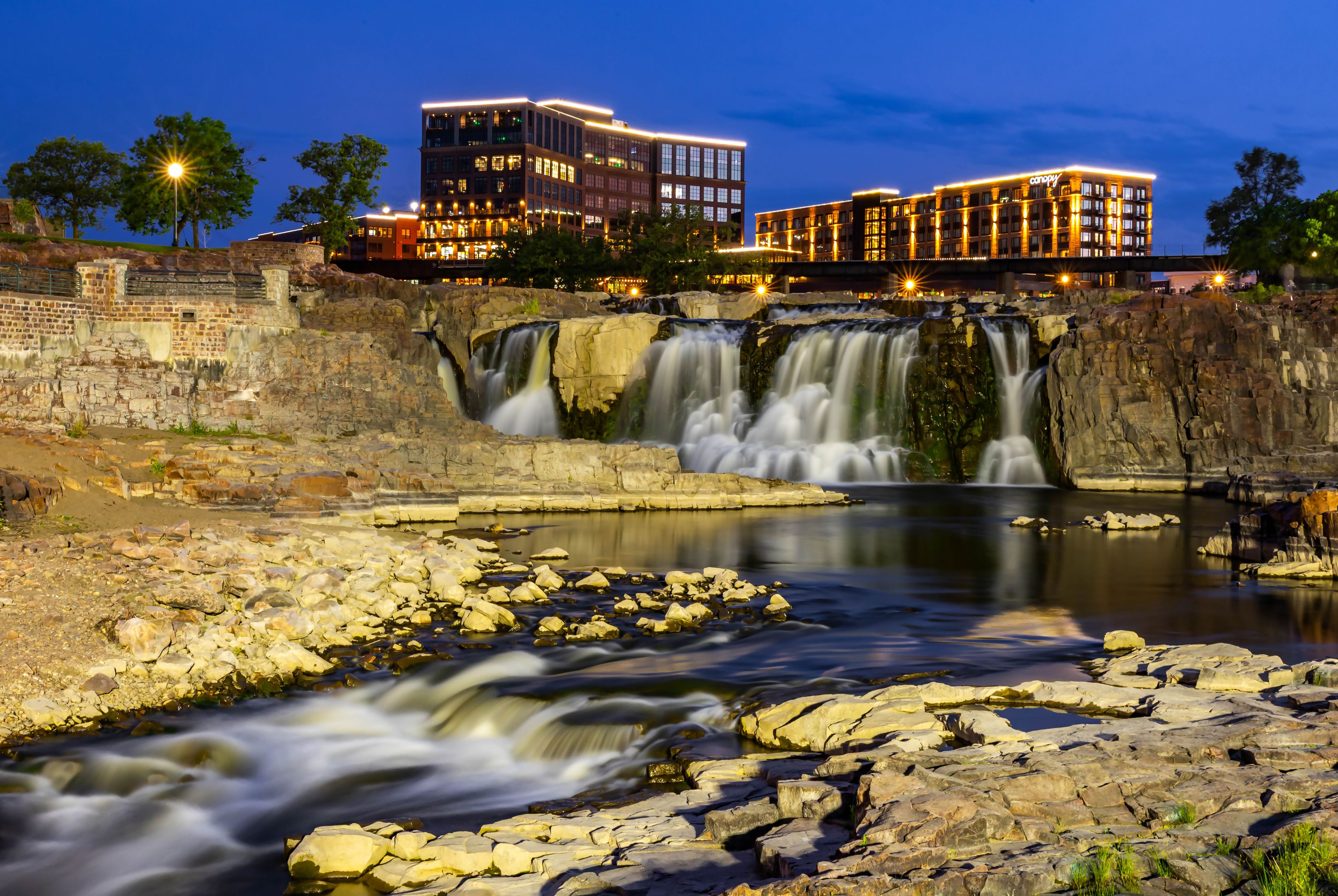 A waterfall cascading over rocks into a river, with illuminated buildings in the background.