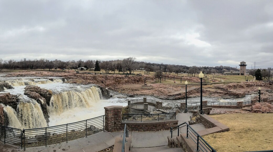 The Big Sioux River flows over rocks in Sioux Falls South Dakota with views of wildlife, ruins, park paths, train track bridge, trees and city in the surrounding area and background