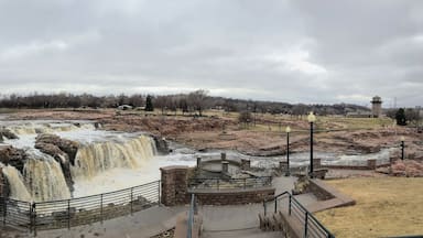 The Big Sioux River flows over rocks in Sioux Falls South Dakota with views of wildlife, ruins, park paths, train track bridge, trees and city in the surrounding area and background