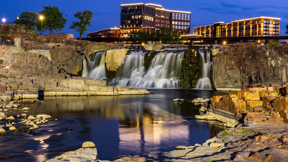 A waterfall cascading over rocky terrain into a reflective pool, with modern buildings illuminated in the background.