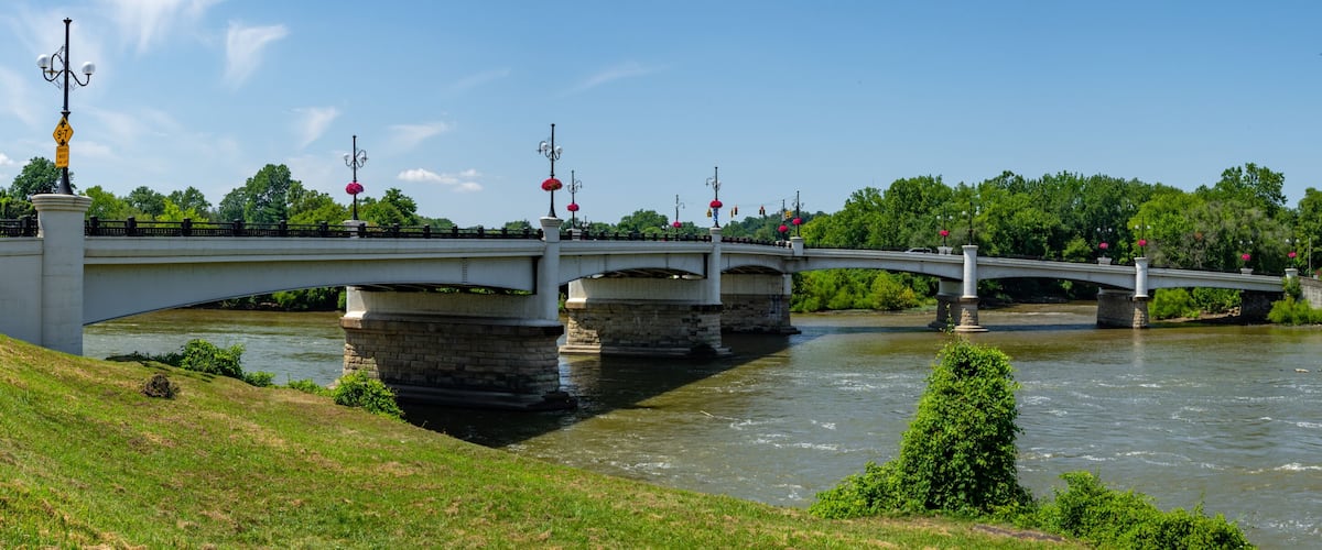 A white bridge over a river with stone pillars, surrounded by greenery and trees.