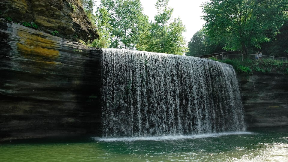 A waterfall cascading over a rocky cliff into a body of water, surrounded by lush greenery and trees.
