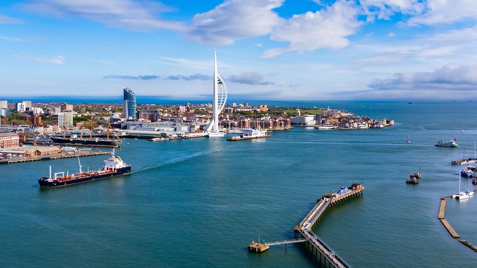 A coastal cityscape with a prominent sail-shaped building, a large cargo ship, and a marina filled with boats.