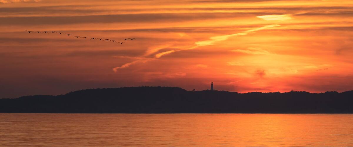 A sunset over a body of water with a silhouette of a landmass and a lighthouse.