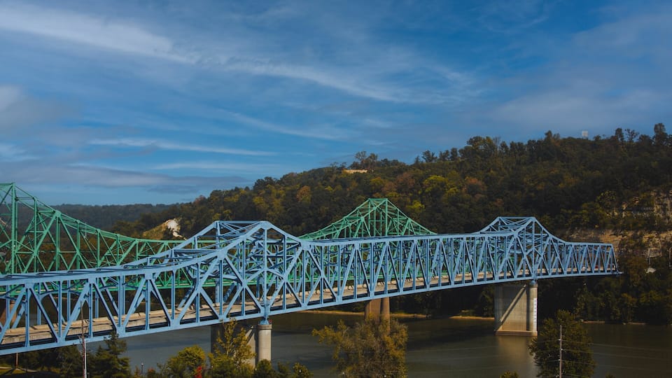 A blue steel bridge spanning a river with a forested hillside in the background.