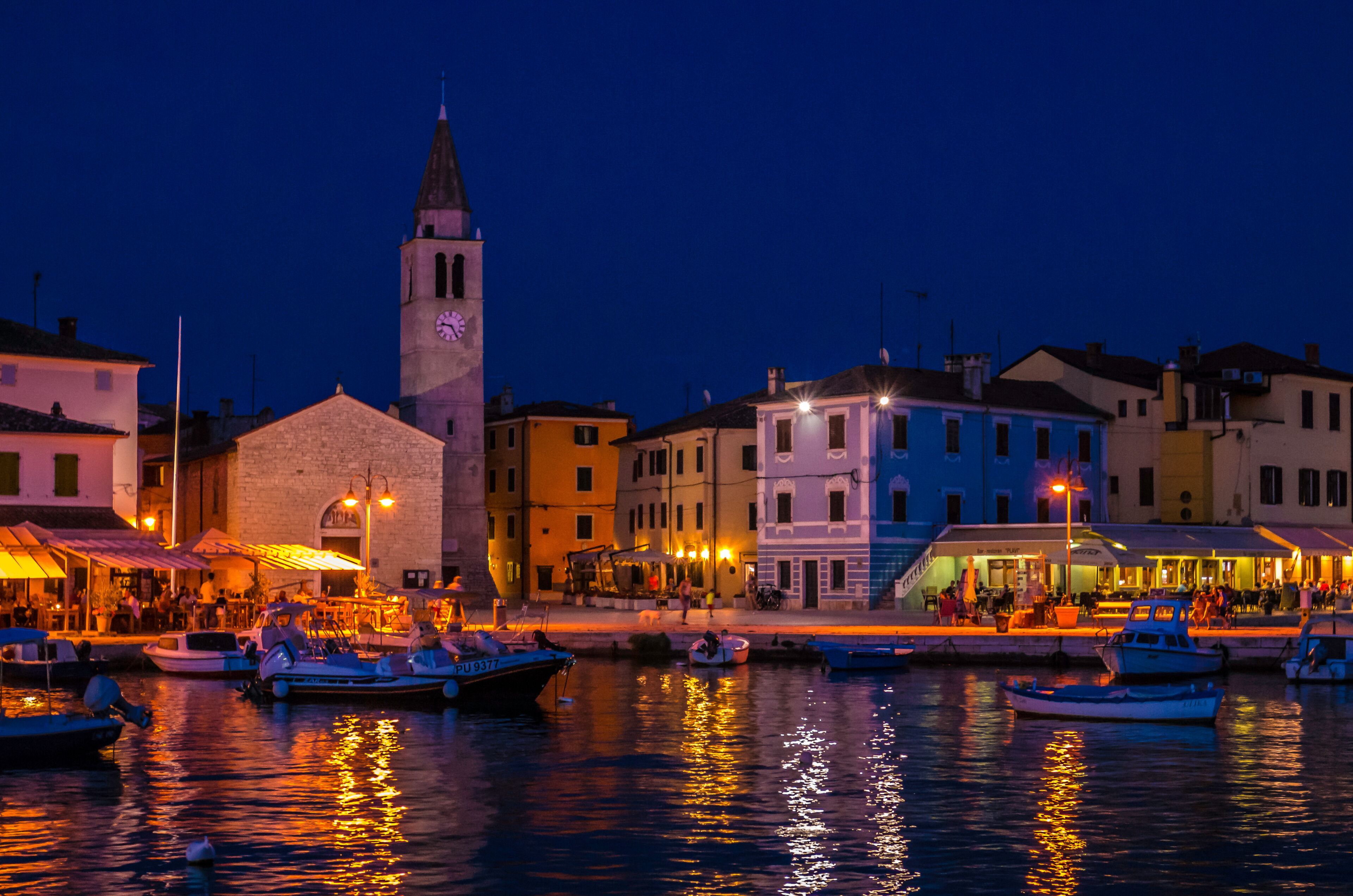 A waterfront town at dusk with colorful buildings, a prominent clock tower, and boats docked along the shore.