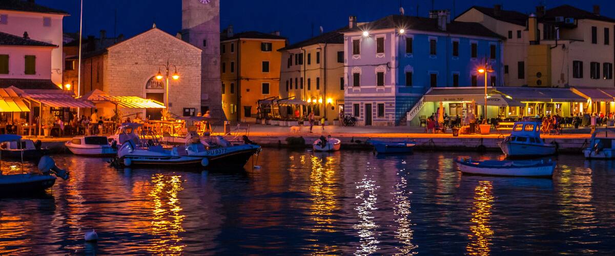 A waterfront town at dusk with colorful buildings, a prominent clock tower, and boats docked along the shore.