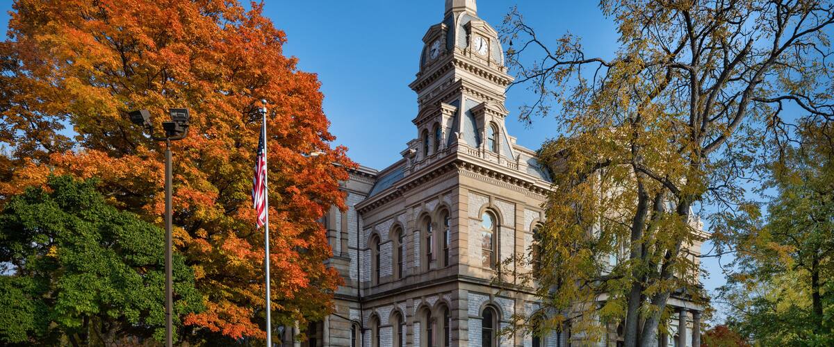 A historic building with a clock tower, surrounded by autumn-colored trees and American flags.