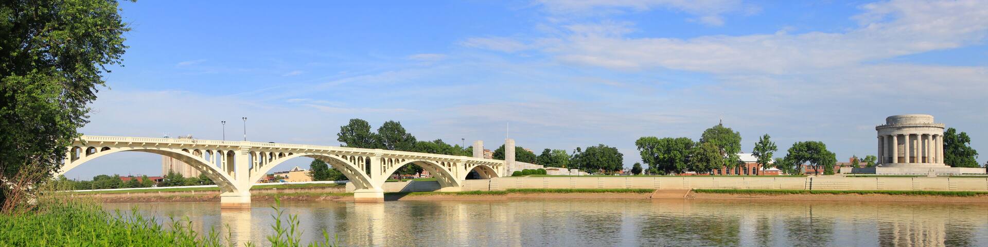 A white bridge over a calm river, surrounded by greenery and a historic monument.