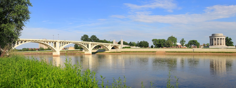 A white bridge over a calm river, surrounded by greenery and a historic monument.