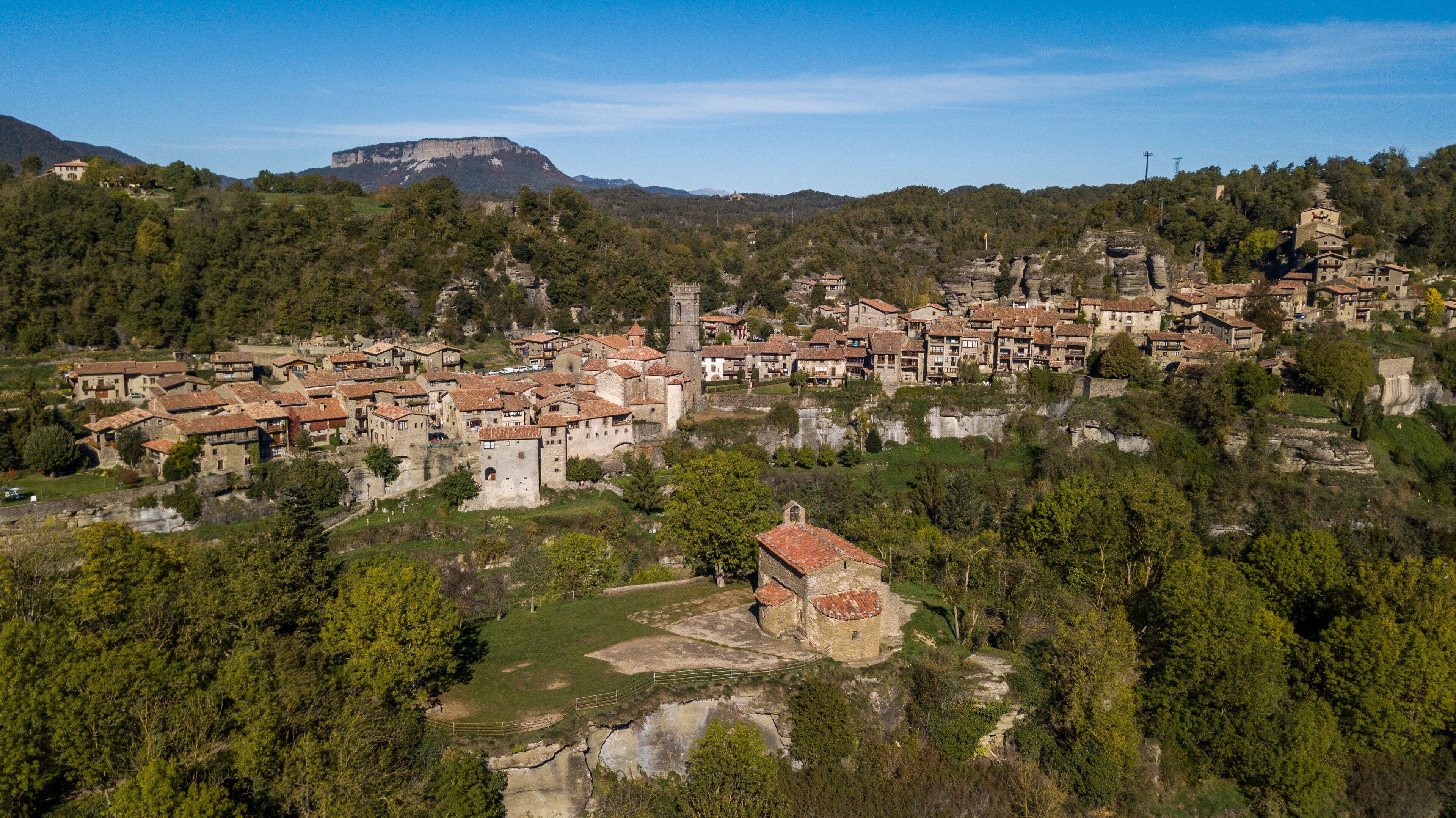 A historic town with terracotta-roofed buildings nestled in a valley, surrounded by dense forests and a prominent mountain in the background.
