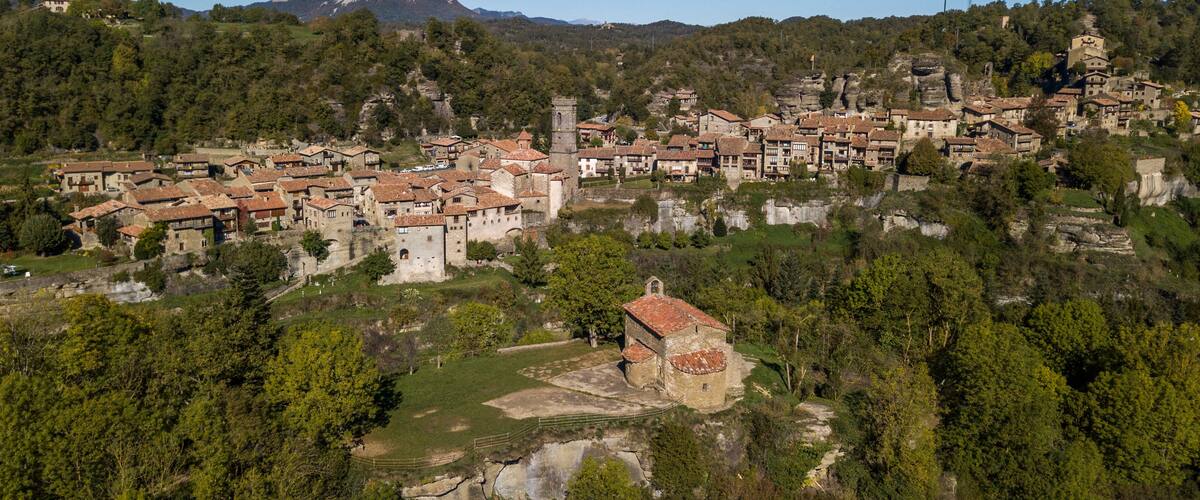A historic town with terracotta-roofed buildings nestled in a valley, surrounded by dense forests and a prominent mountain in the background.