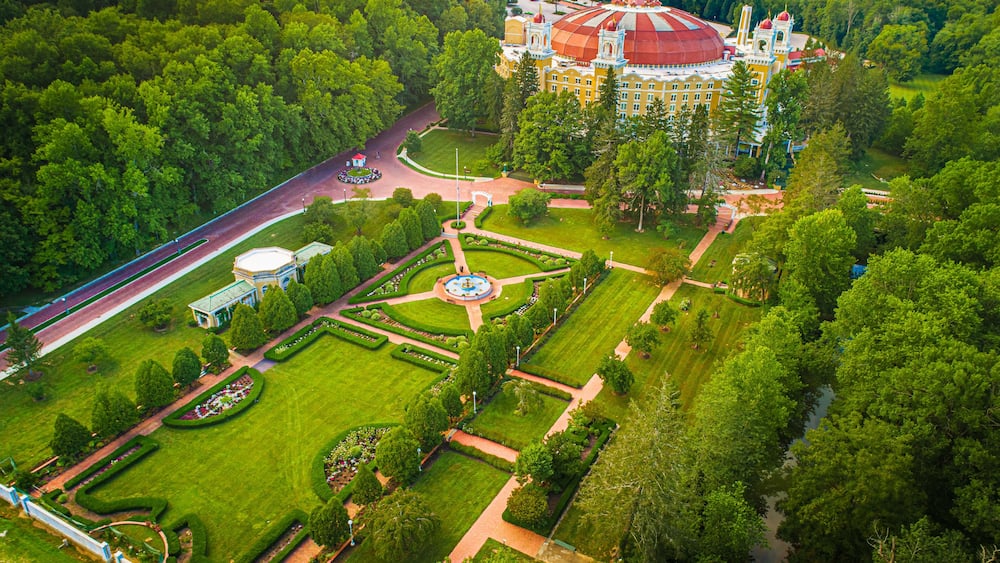 Une vue aérienne d’un parc avec des pelouses bien entretenues, une fontaine centrale et un grand bâtiment surmonté d’un dôme rouge.