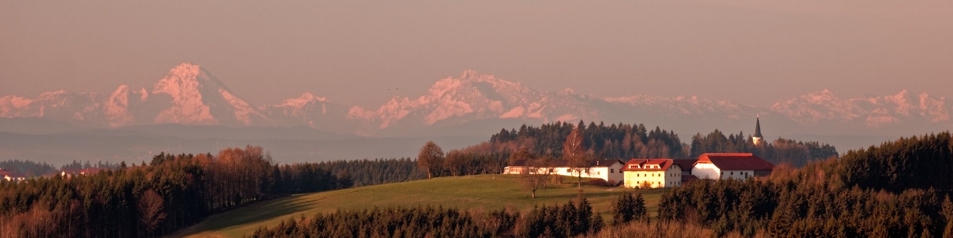 A rural landscape with a church, houses, and a mountain range in the background.