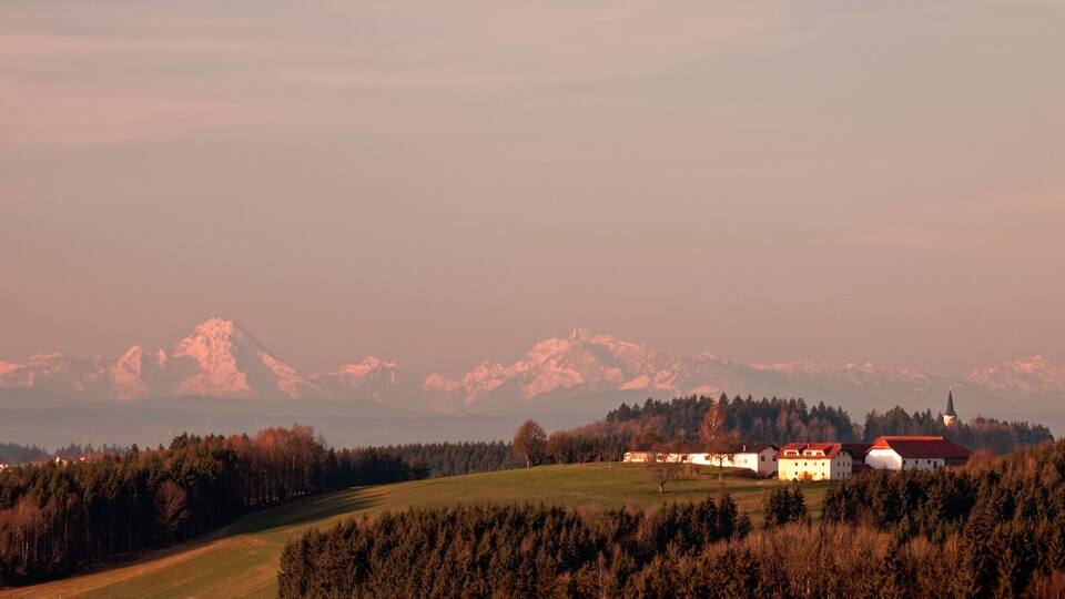 A rural landscape with a church, a large white building with a red roof, and snow-capped mountains in the background.