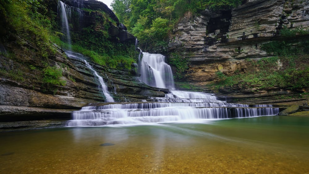 A multi-tiered waterfall cascading into a clear pool surrounded by rocky cliffs and lush greenery.