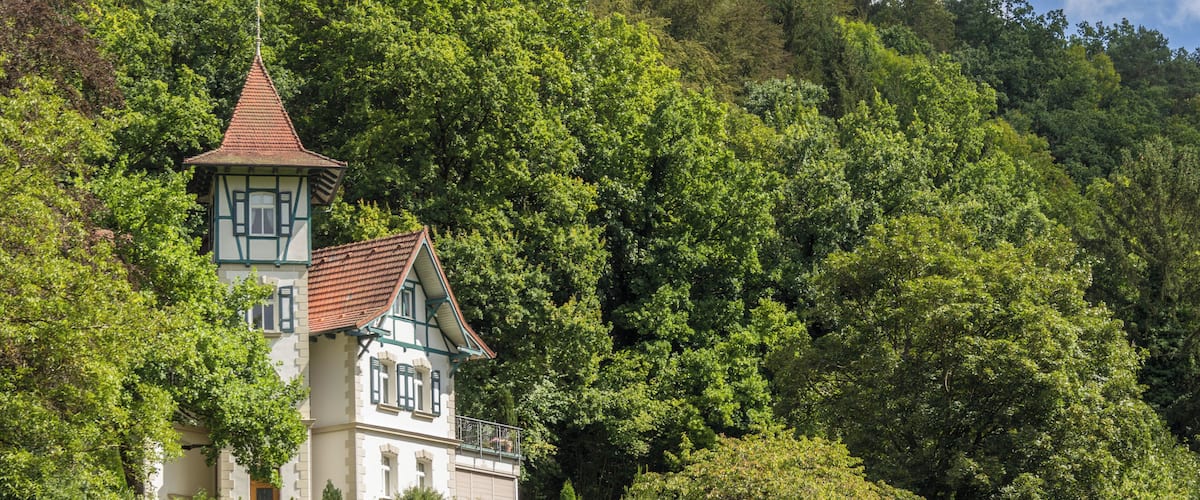 A white building with a red roof and a tower, surrounded by lush green trees on a hillside.