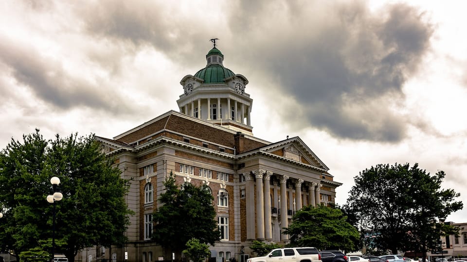 A large, historic building with a prominent dome and columns, surrounded by trees and parked cars.