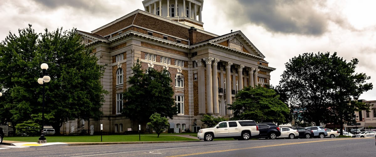 A large, historic building with a prominent dome and columns, surrounded by trees and parked cars.