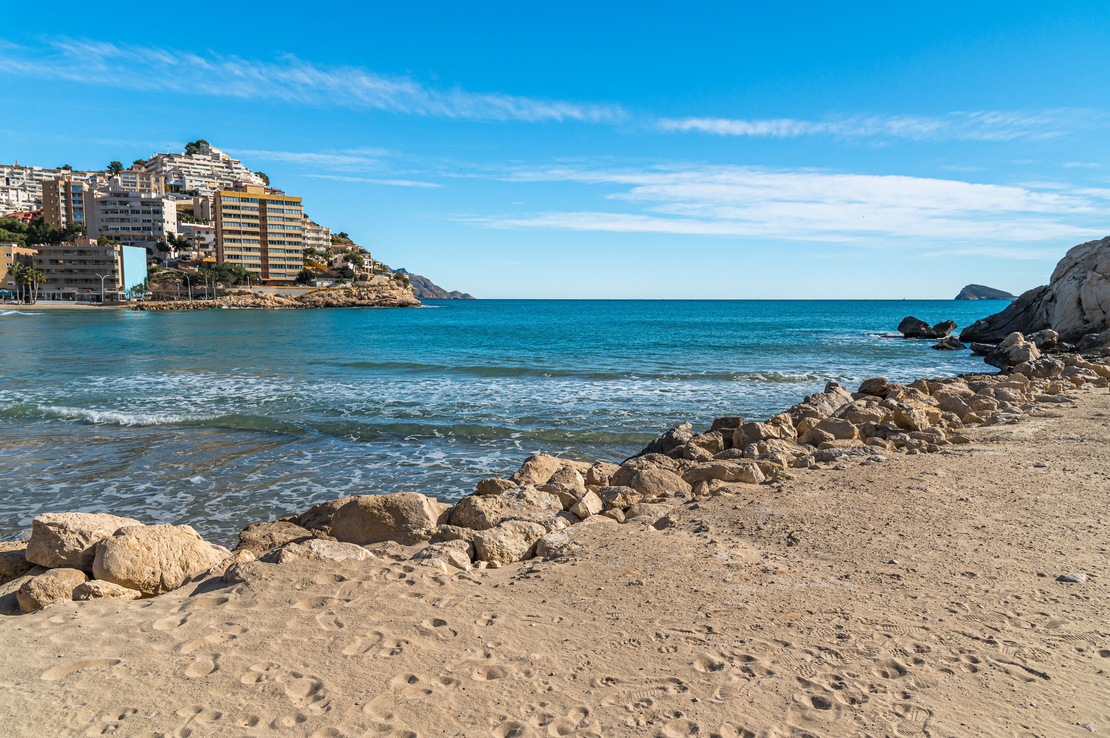 A sandy beach with rocky outcrops and a clear blue sea. In the background, there are buildings on a hillside.