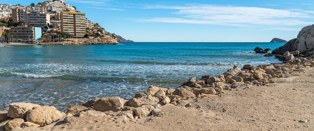 A sandy beach with rocky outcrops and a clear blue sea. In the background, there are buildings on a hillside.