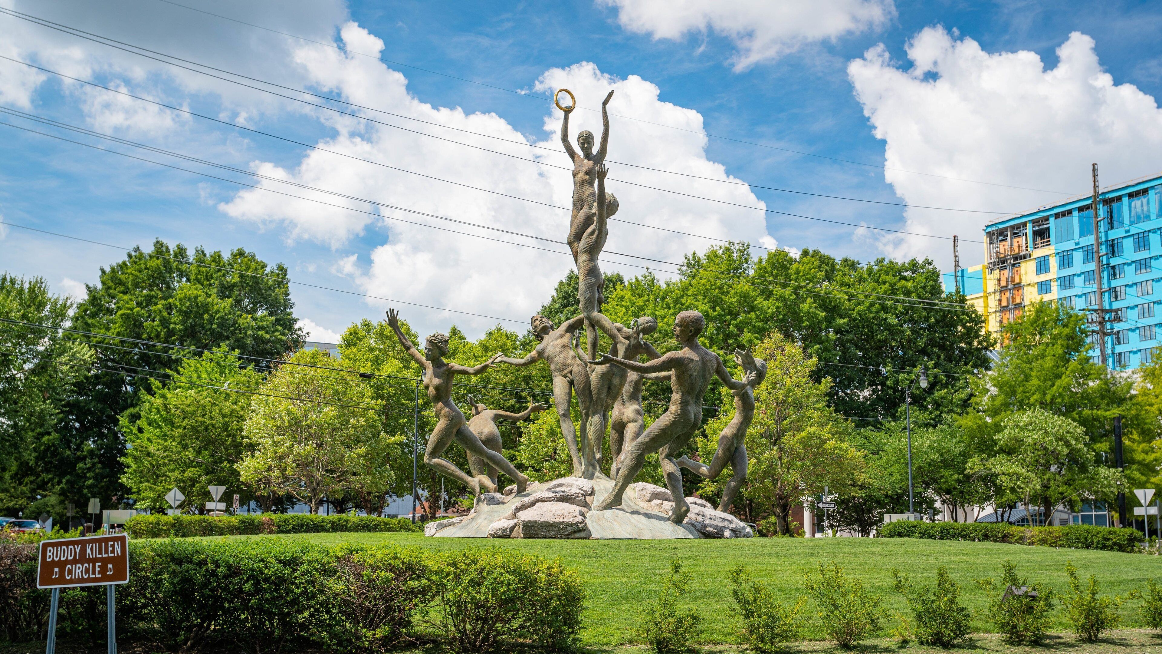 A bronze sculpture of a woman holding a ring, surrounded by figures in a park with trees and a building in the background.