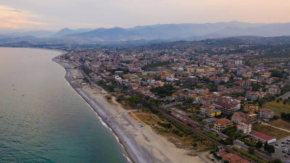 An aerial view of a coastal city with a beach, residential areas, and mountains in the background.