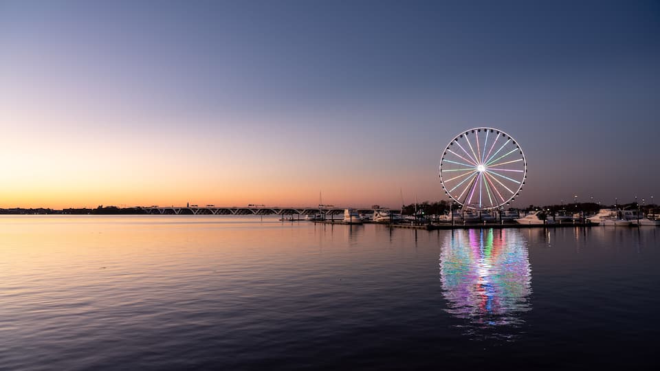 A large Ferris wheel reflecting on calm water at sunset.