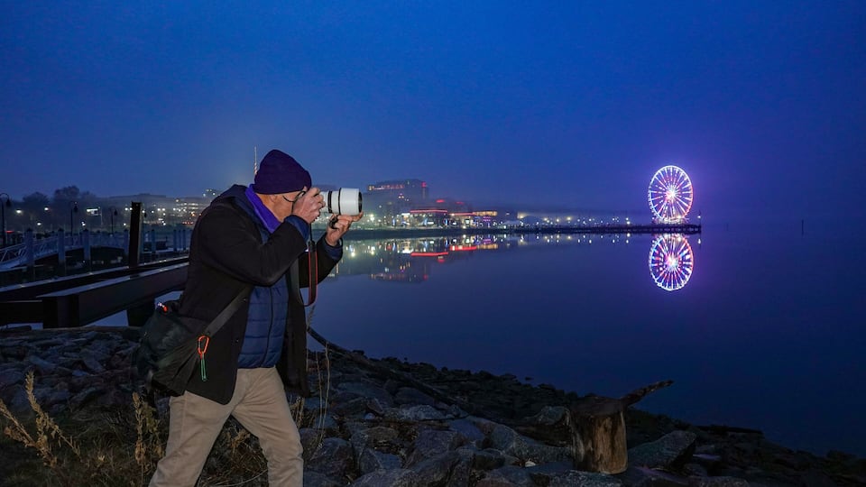A person taking a photo with a camera, a body of water, and a brightly lit Ferris wheel in the background.