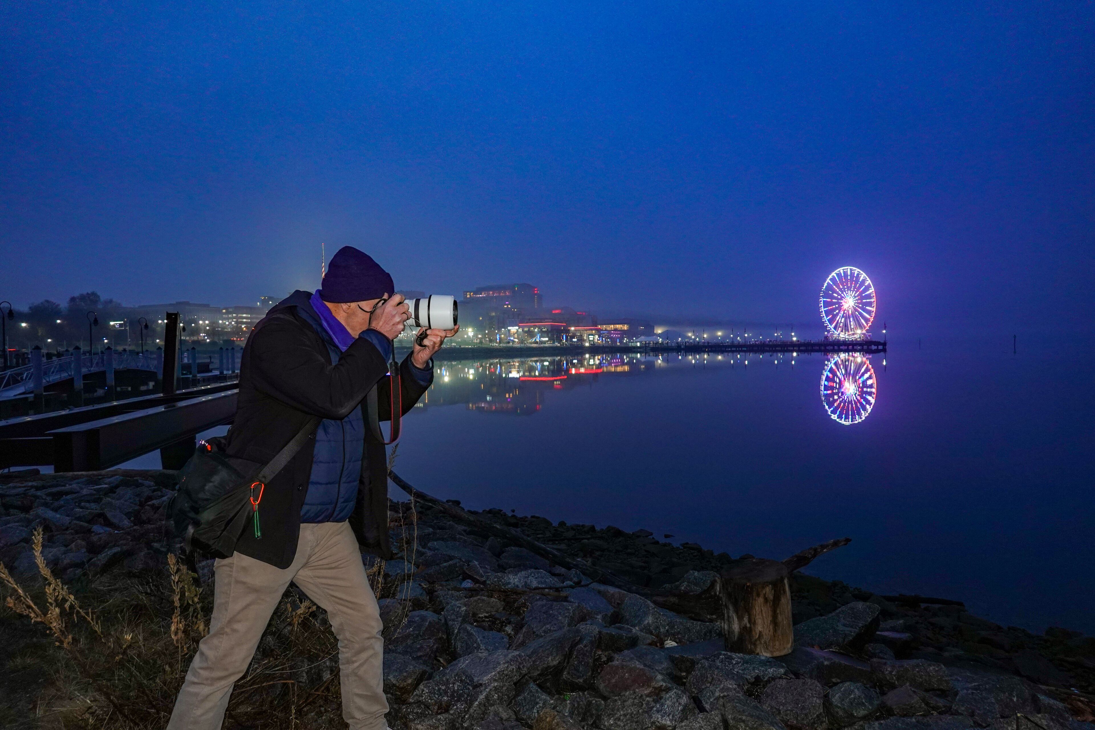 A person taking a photo with a camera, a body of water, and a brightly lit Ferris wheel in the background.