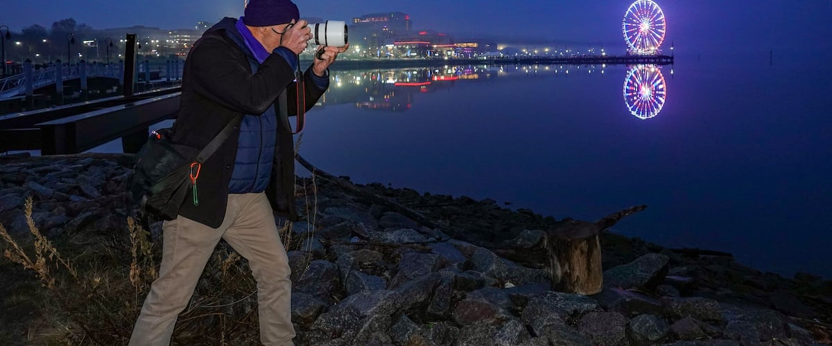 A person taking a photo with a camera, a body of water, and a brightly lit Ferris wheel in the background.