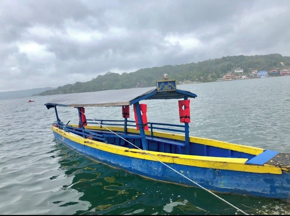 A blue and yellow boat with a canopy and life jackets on the side, docked on calm water with a cloudy sky and a green hillside in the background.