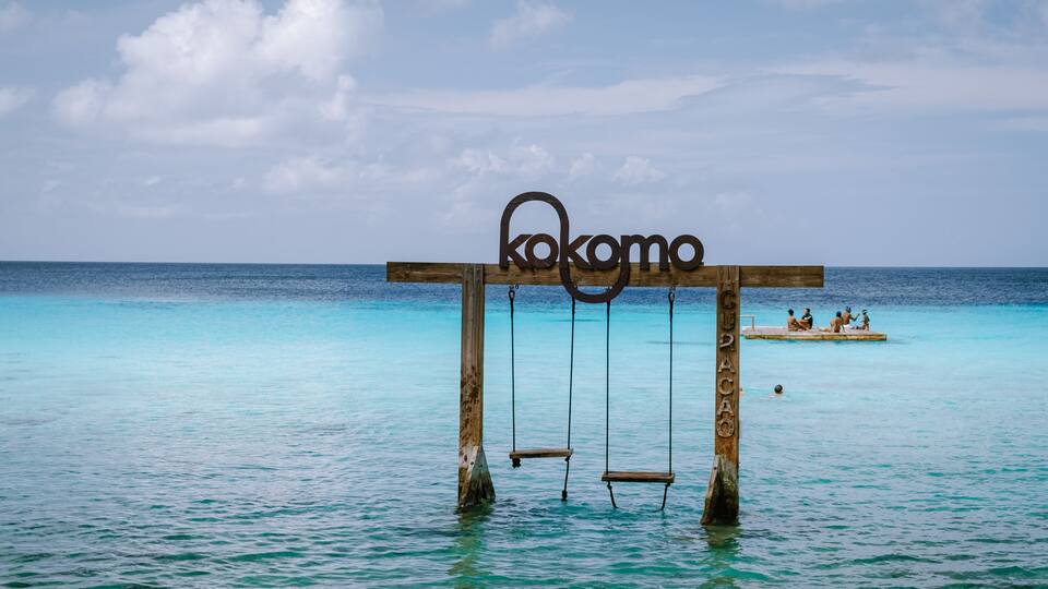 A wooden swing set with the word 'kokomo' in the water, a clear blue sea, and a small island with people in the background.