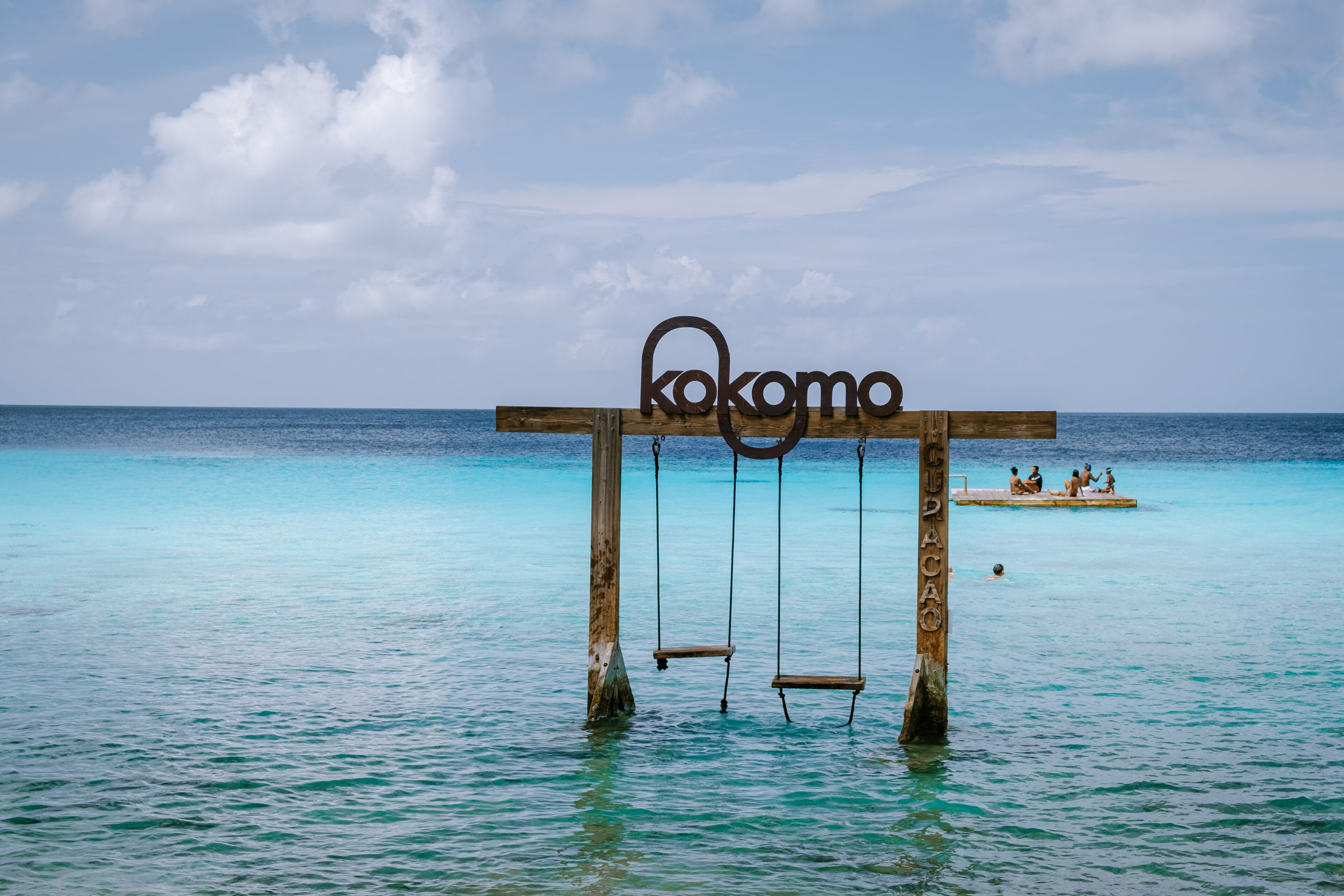 A wooden swing set with the word 'kokomo' in the water, a clear blue sea, and a small island with people in the background.