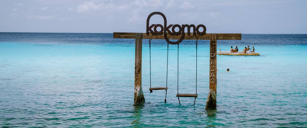 A wooden swing set with the word 'kokomo' in the water, a clear blue sea, and a small island with people in the background.