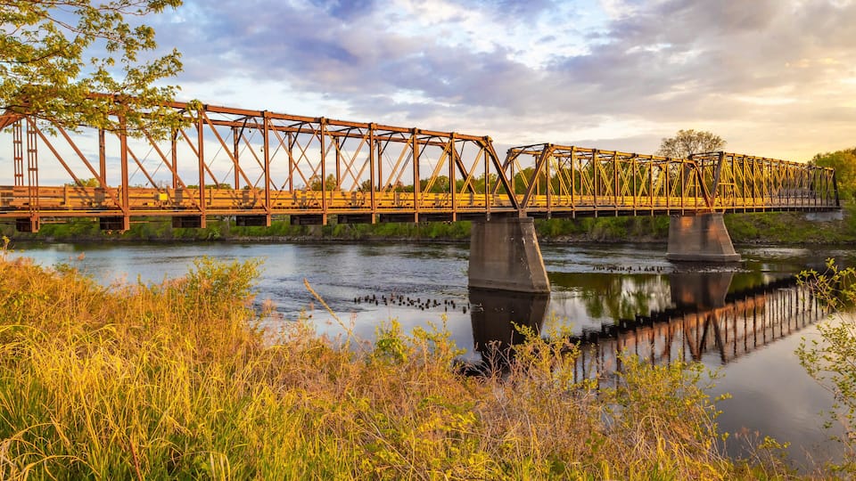 A steel bridge over a calm river with surrounding greenery and a cloudy sky.