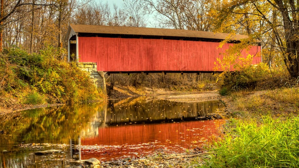 A red covered bridge over a calm river with autumn foliage.