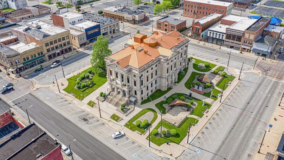 An aerial view of a city with a prominent historic building with a red roof, surrounded by green spaces and a wide street.
